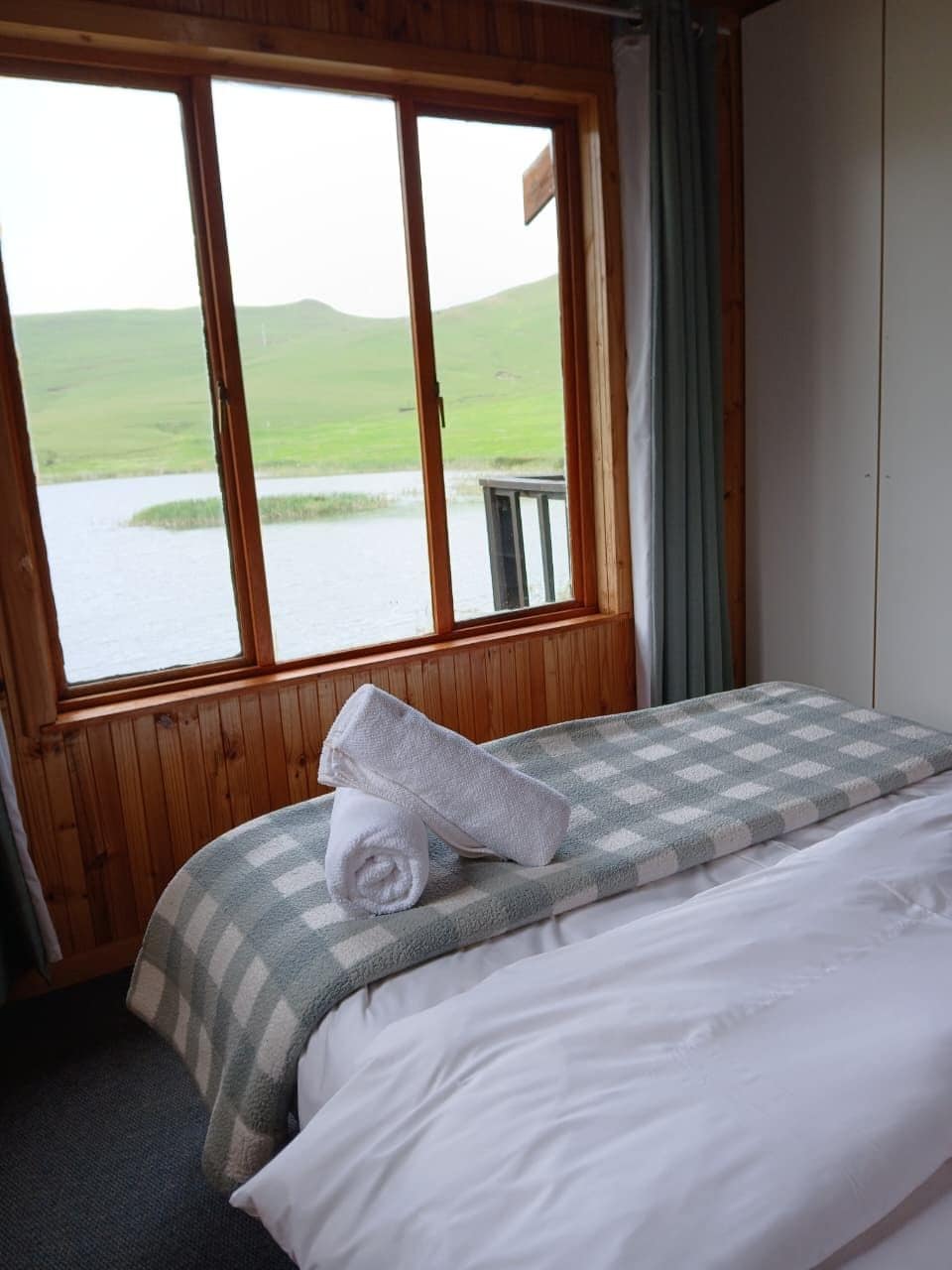 A room with a bed, a window with a view of a green landscape and water, and wooden walls.