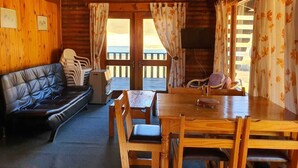 Ein gemütliches Blockhäuschen im Inneren mit Holzausstattung, einer Couch, einem Esstisch mit Stühlen und Blick durch die Tür.