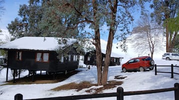 A wooden cabin with a snow-covered roof, surrounded by trees and a wooden fence.