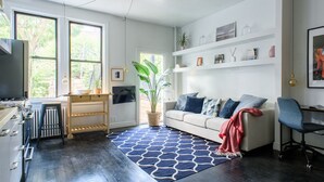 Una sala de estar moderna con un sofá, una mesa de centro y un televisor. Cuenta con una zona de cocina con estufa y armarios. La habitación tiene ventanas grandes y una planta en maceta.