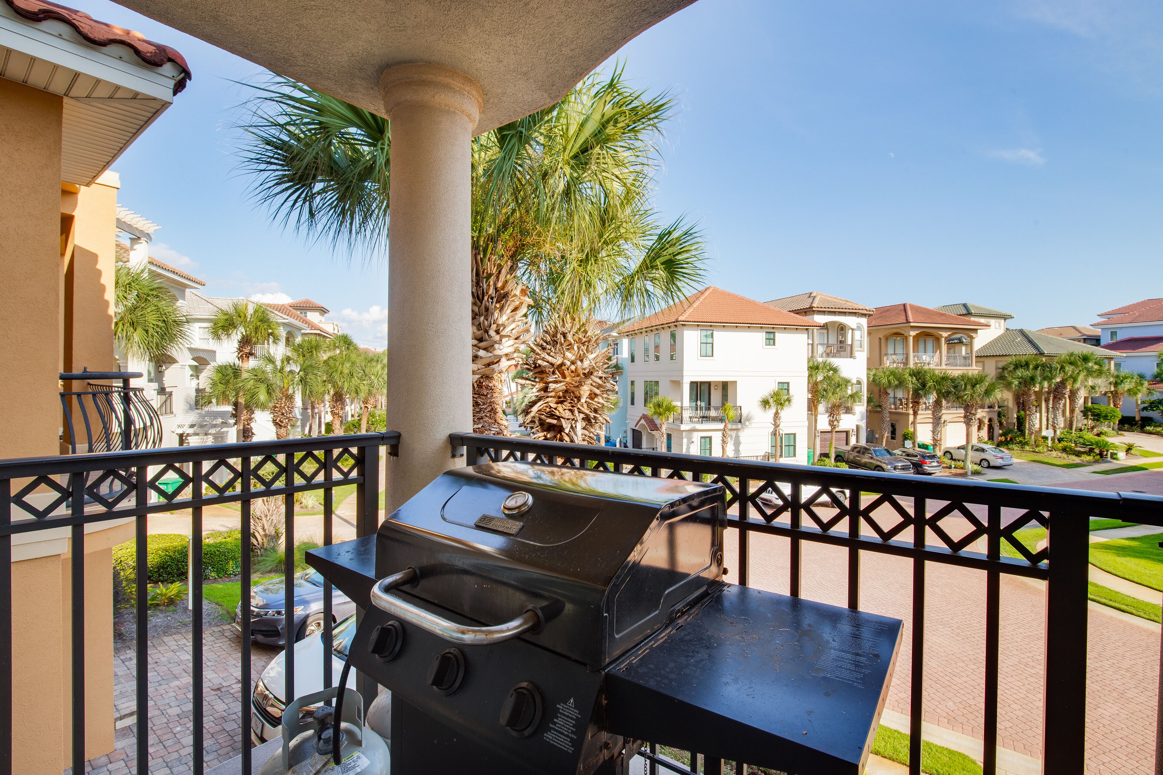 A balcony with a grill, palm trees, and a view of a residential area with houses and a street.