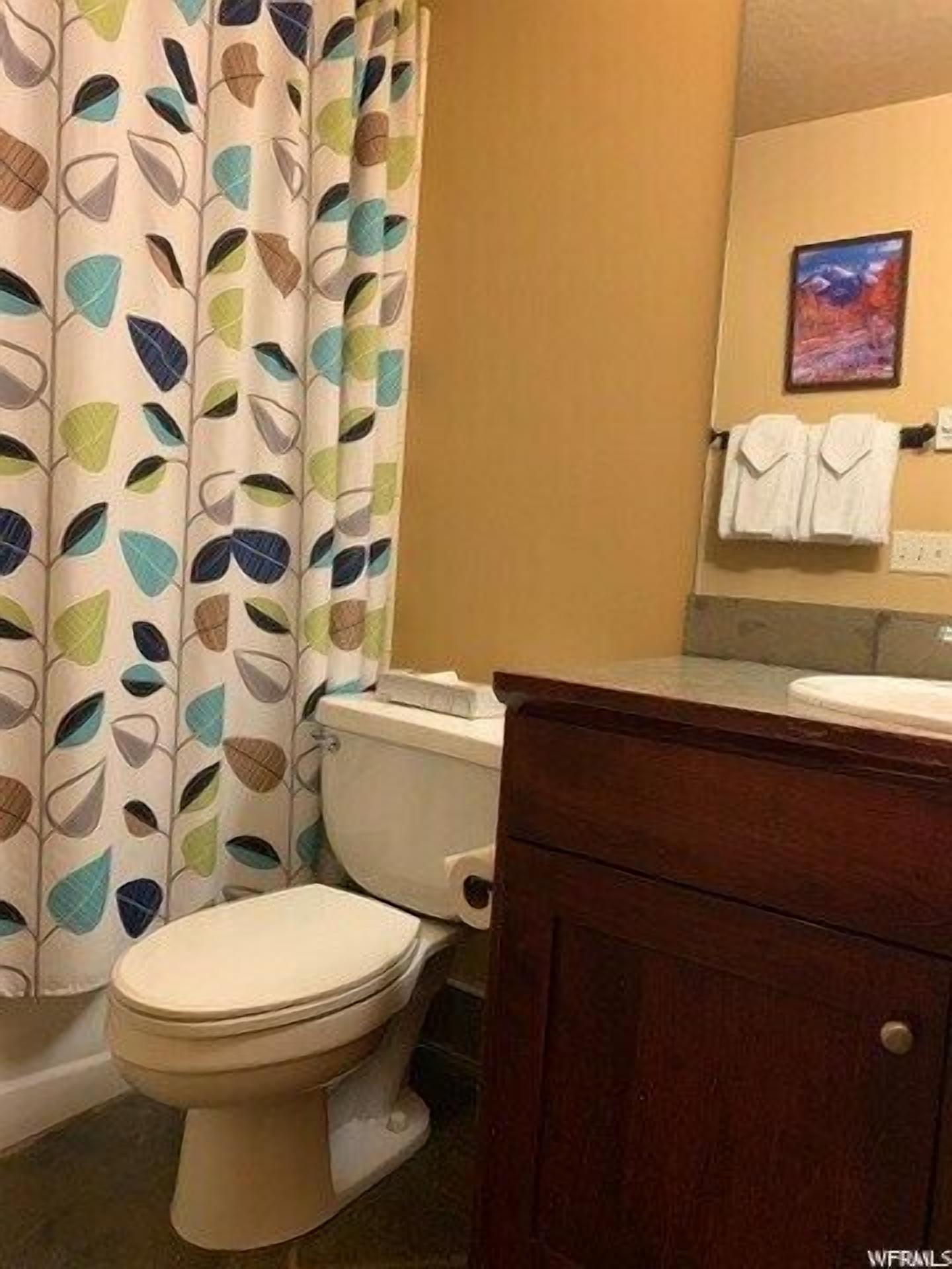 A bathroom with a toilet, a sink, a mirror, and a shower curtain with a leaf pattern.