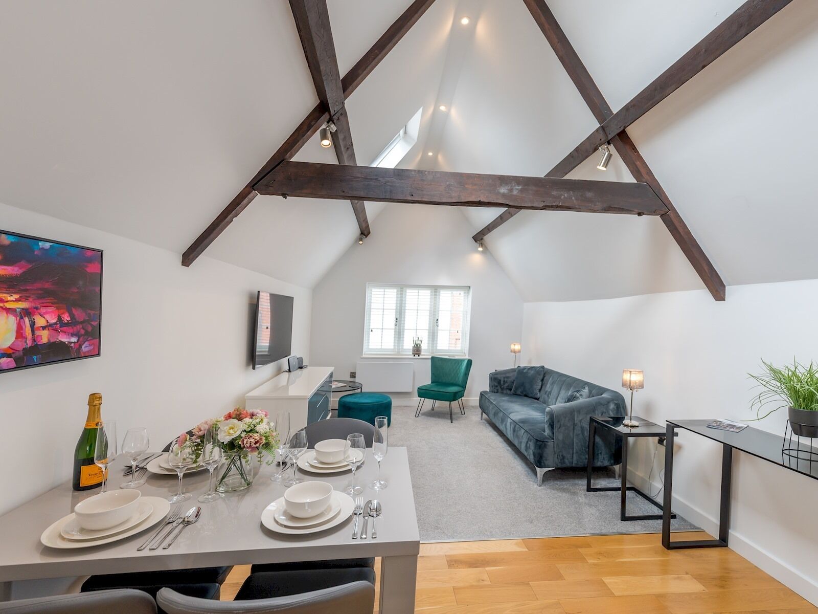 A modern loft apartment with exposed wooden beams, a dining area with a table set for four, a sofa, and a TV mounted on the wall.