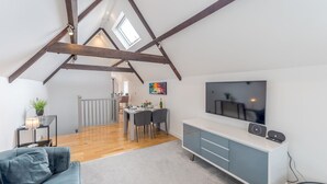 En moderne loftlejlighed med synlige træbjælker, et spiseområde, en stue med sofa og fjernsyn, samt et køkken i baggrunden.