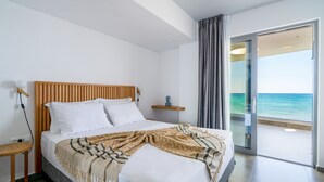 Ein modernes Schlafzimmer mit einem Holzkopfteil, einem Bett mit weißer Bettwäsche, einer Tagesdecke, einem Nachttisch und Meerblick durch eine Glastür.