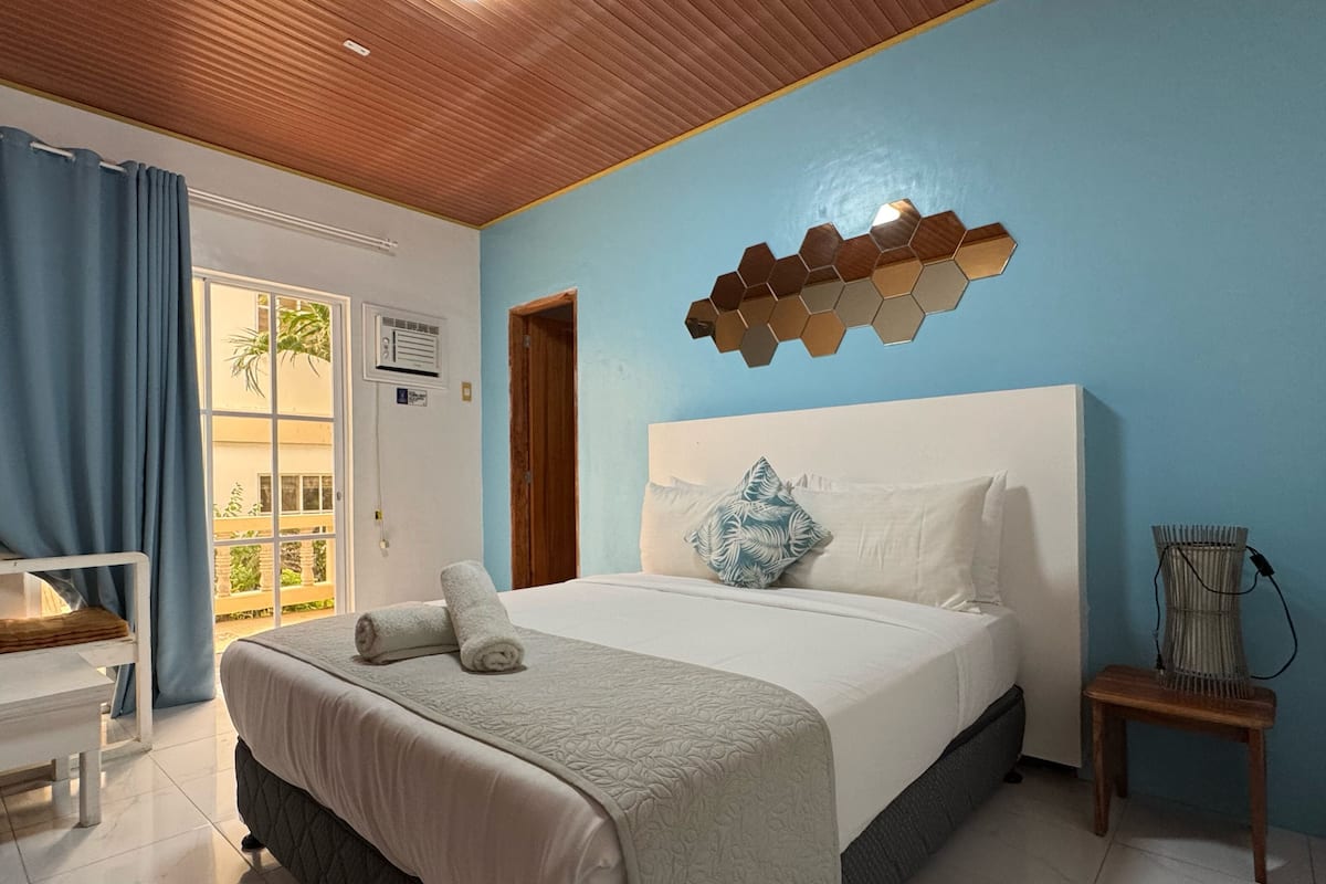 A bedroom with a bed, a desk, a chair, a wall-mounted light fixture, and a view of the outdoors.