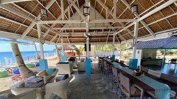 Un espacio de estar al aire libre con mesas y sillas, un ventilador y vistas a la playa y al océano.