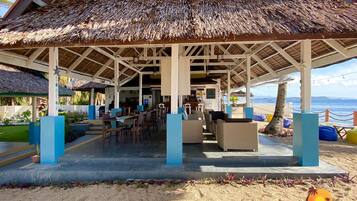A beachside lounge with thatched roofs, outdoor seating, and a view of the ocean.