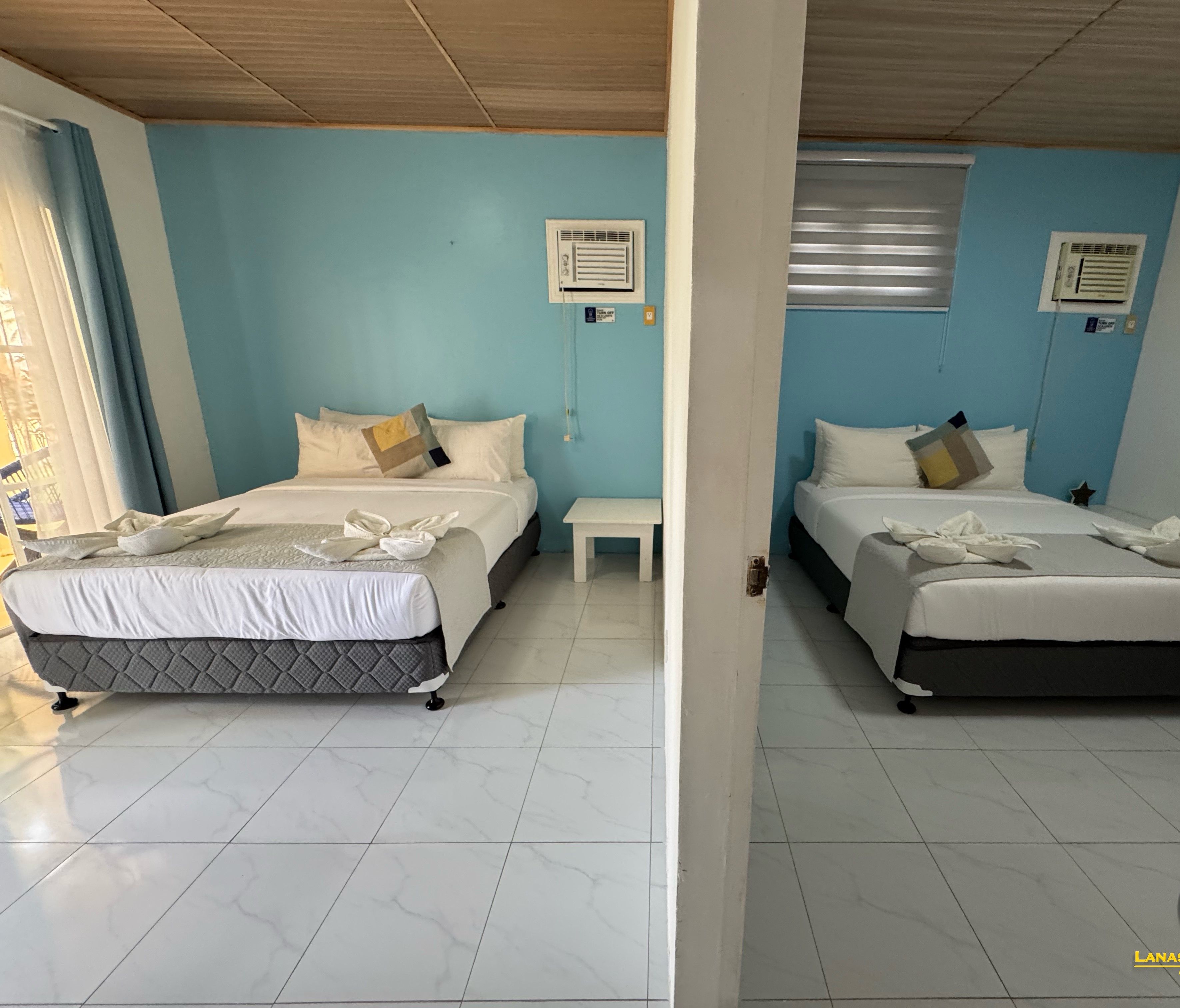 Dos camas gemelas idénticas con ropa blanca de cama y una mesita de noche pequeña en una habitación con paredes azul claro y pisos de baldosas blancas.
