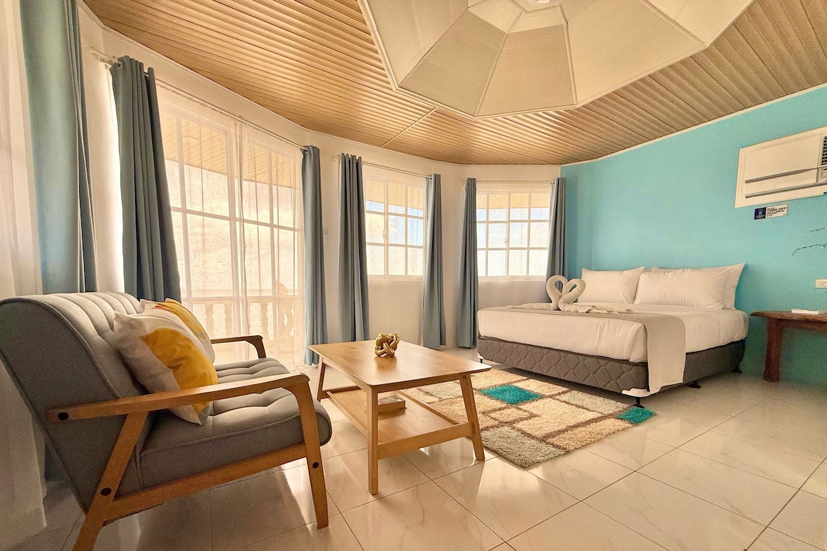 A modern hotel room with a bed, a sofa, a wooden table, and large windows with curtains.