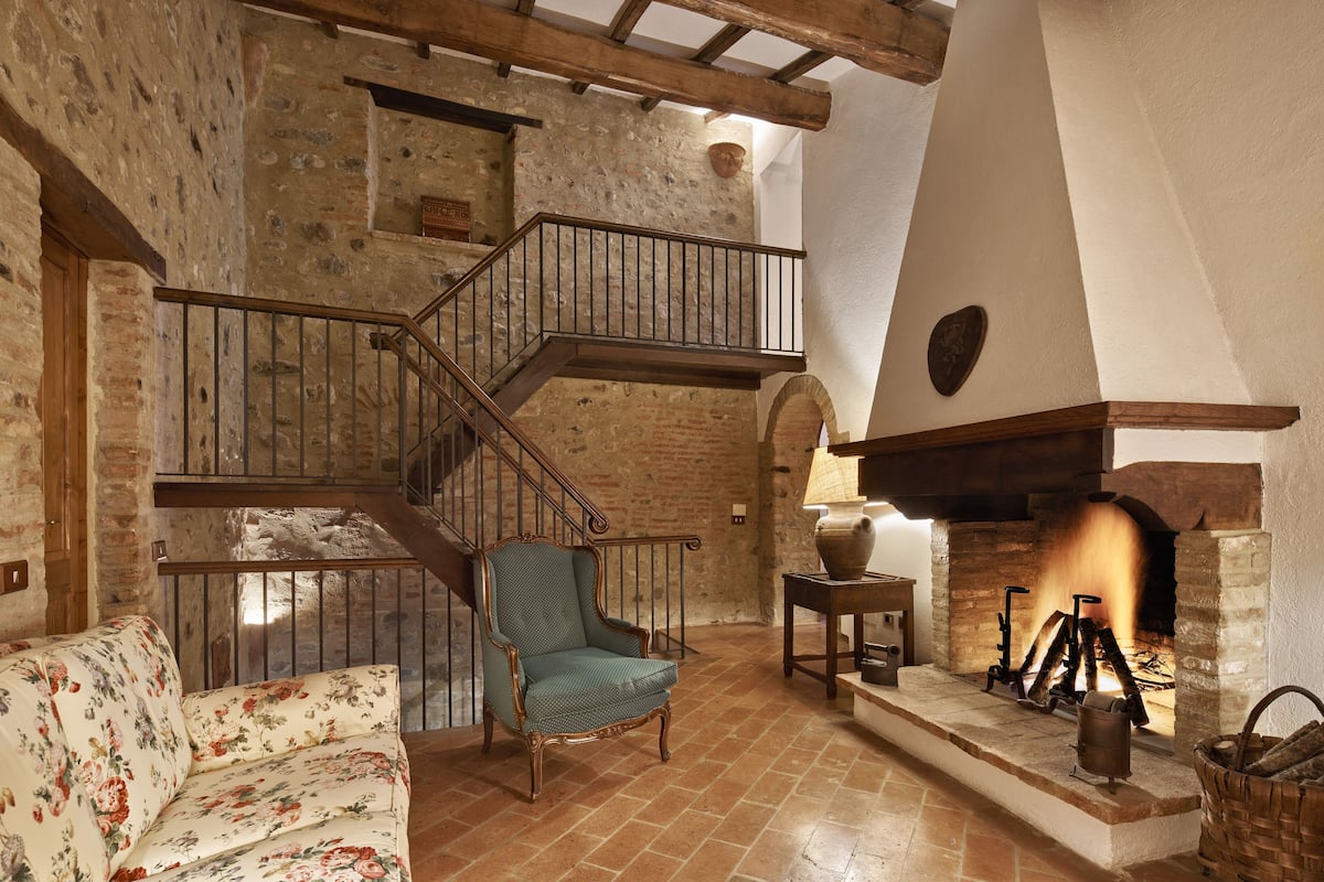 A rustic interior with a stone wall, a wooden staircase, a floral sofa, a vintage armchair, and a fireplace with logs.