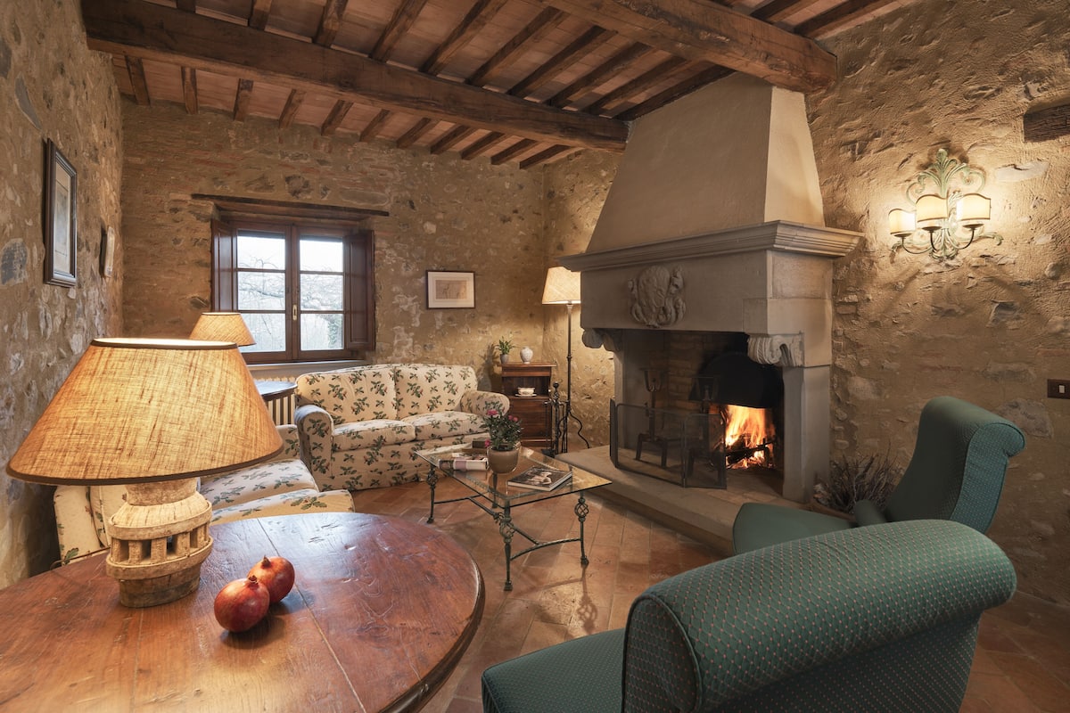 A rustic living room with a stone wall, wooden beams, a fireplace, a sofa, a table with a lamp, and a pomegranate on the table.
