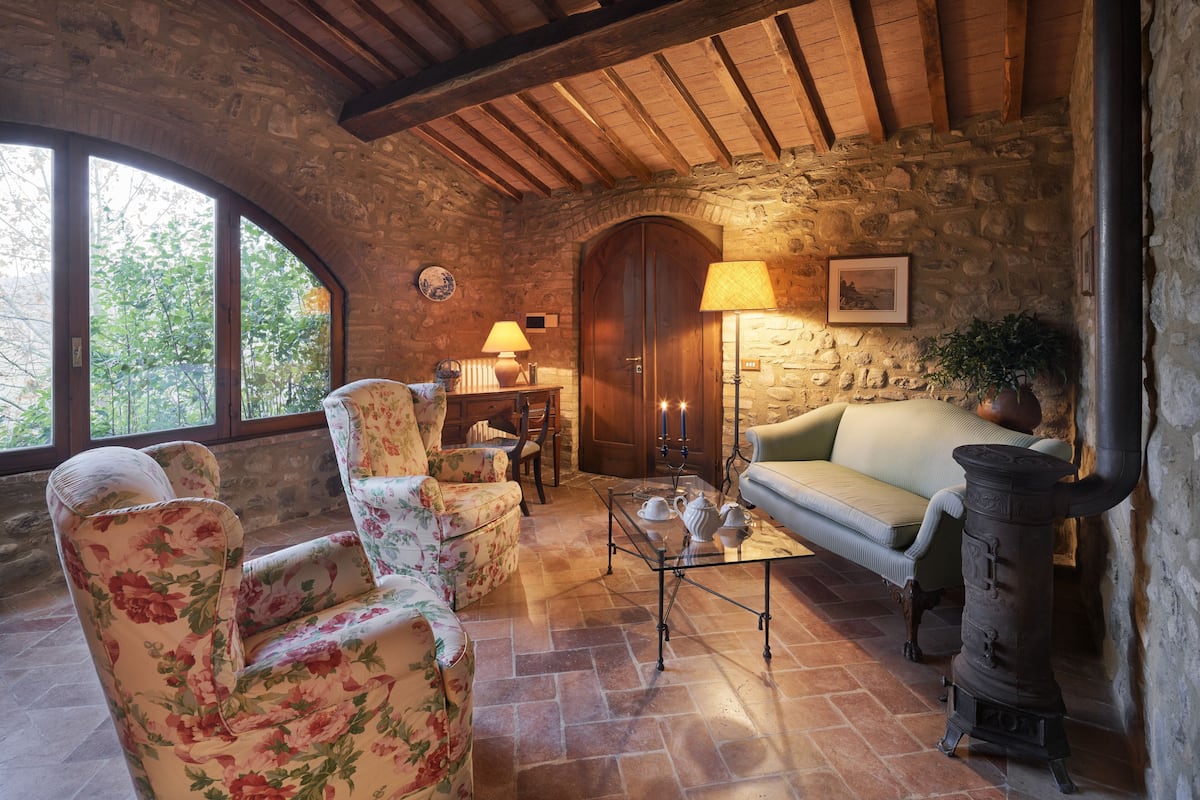 A rustic room with stone walls, wooden beams, a stone fireplace, floral armchairs, a sofa, and a table with tea set.
