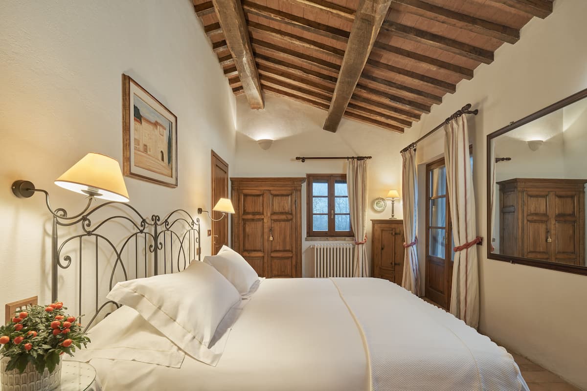 A bedroom with a large bed, wooden ceiling beams, a window with curtains, and a mirror on the wall.