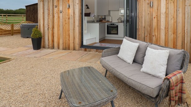 A cozy outdoor seating area with a sofa and a table, a wooden exterior, and an open kitchen visible through a glass door. - Hillside Hut (Ashbourne)
