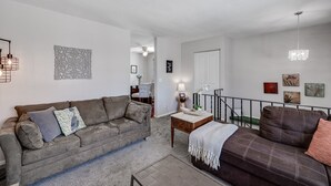 Una sala de estar con un sofá gris, una mesita de madera y un sofá marrón. Se ve una escalera que conduce a otra habitación.