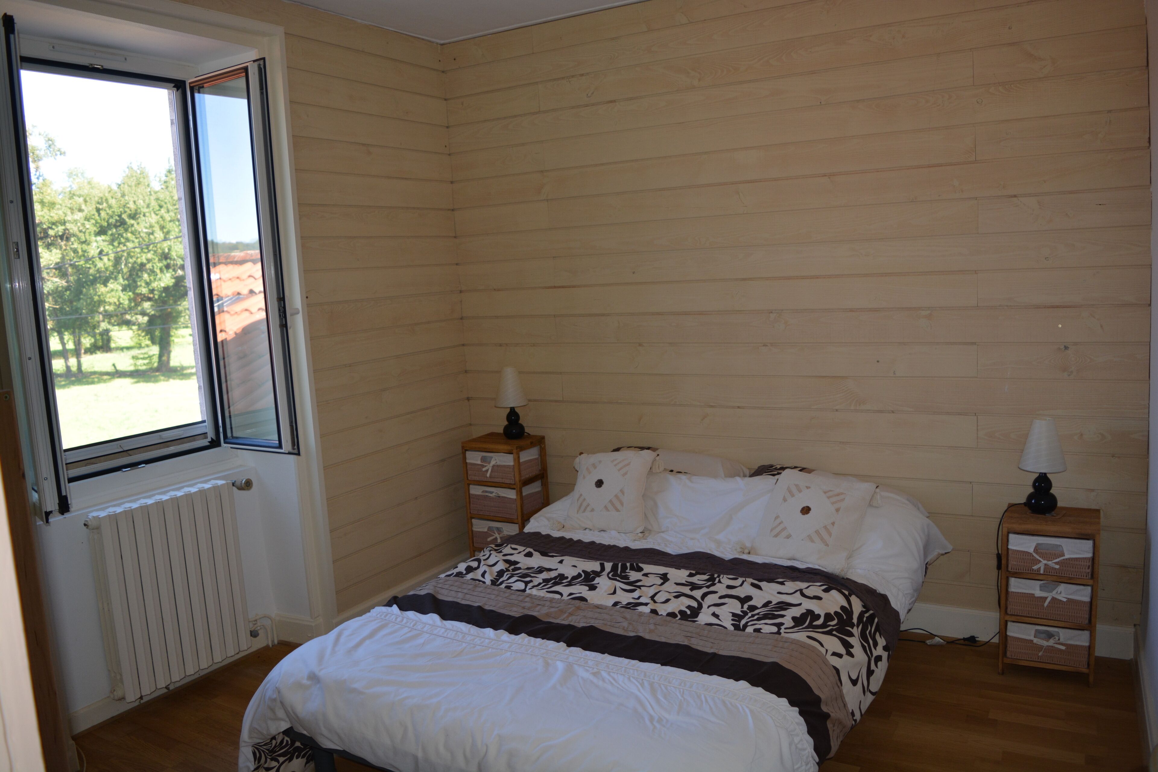 A bedroom with a wooden paneled wall, a large window, a bed with white bedding, two bedside tables with lamps, and a radiator.
