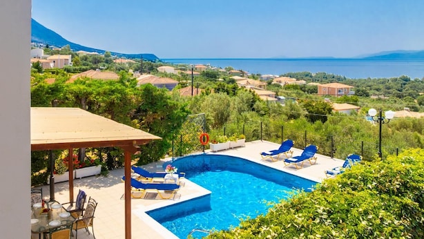 A pool area with lounge chairs and a pergola, overlooking a coastal landscape with houses and a clear sky.