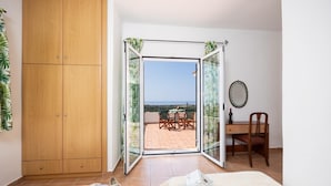 A bedroom with a wooden wardrobe, a bed, a desk with a chair, and a sliding glass door leading to an outdoor patio with a table and chairs.