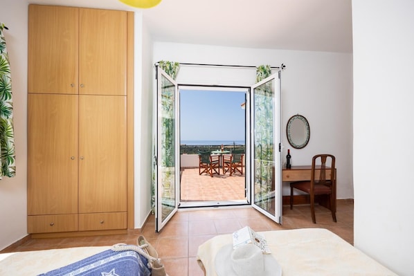 Ein Schlafzimmer mit einem Holzschrank, einem Bett, einem Schreibtisch mit Stuhl und einer Glastür zum Außenbereich mit Tisch und Stühlen.