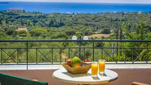 A table with a fruit bowl and two glasses of orange juice, overlooking a scenic view of the sea and greenery.