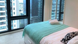 Un dormitorio con un ventanal grande, una cama con cubrecama verde y blanco, y un almohadón blanco.