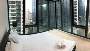 Una habitación de hotel moderna con una cama grande, vistas de la ciudad a través de ventanas de techo a piso, y un paisaje urbano visible desde el exterior.