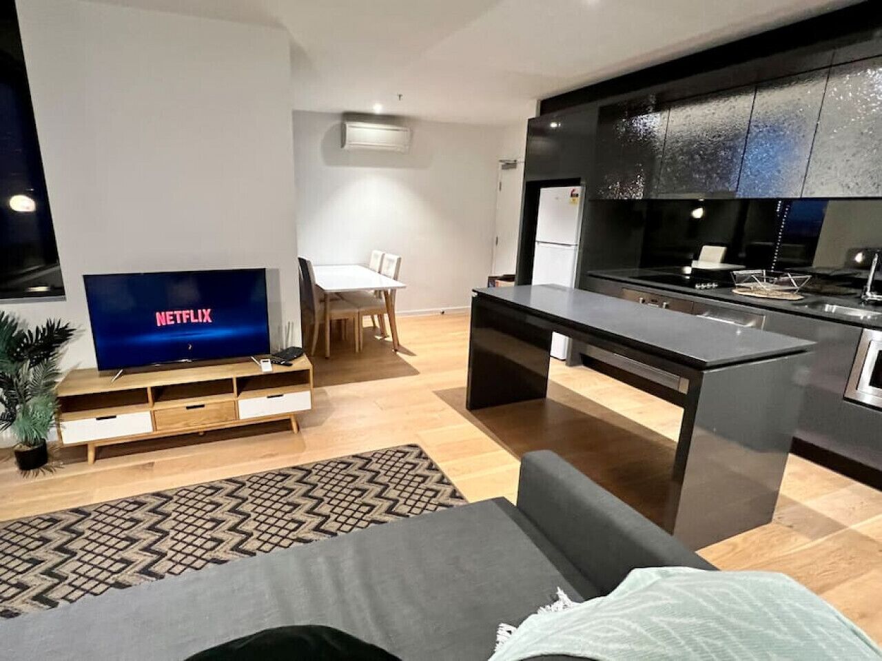 Een moderne woonkamer met een flatscreen televisie, een bank, een eethoek en een keuken met donkere kastjes.