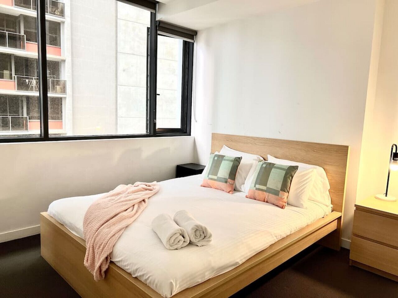 Uma cama bem arrumada com travesseiros e um edredom, uma mesa de cabeceira com abajur, e uma janela com vista para um edifício.