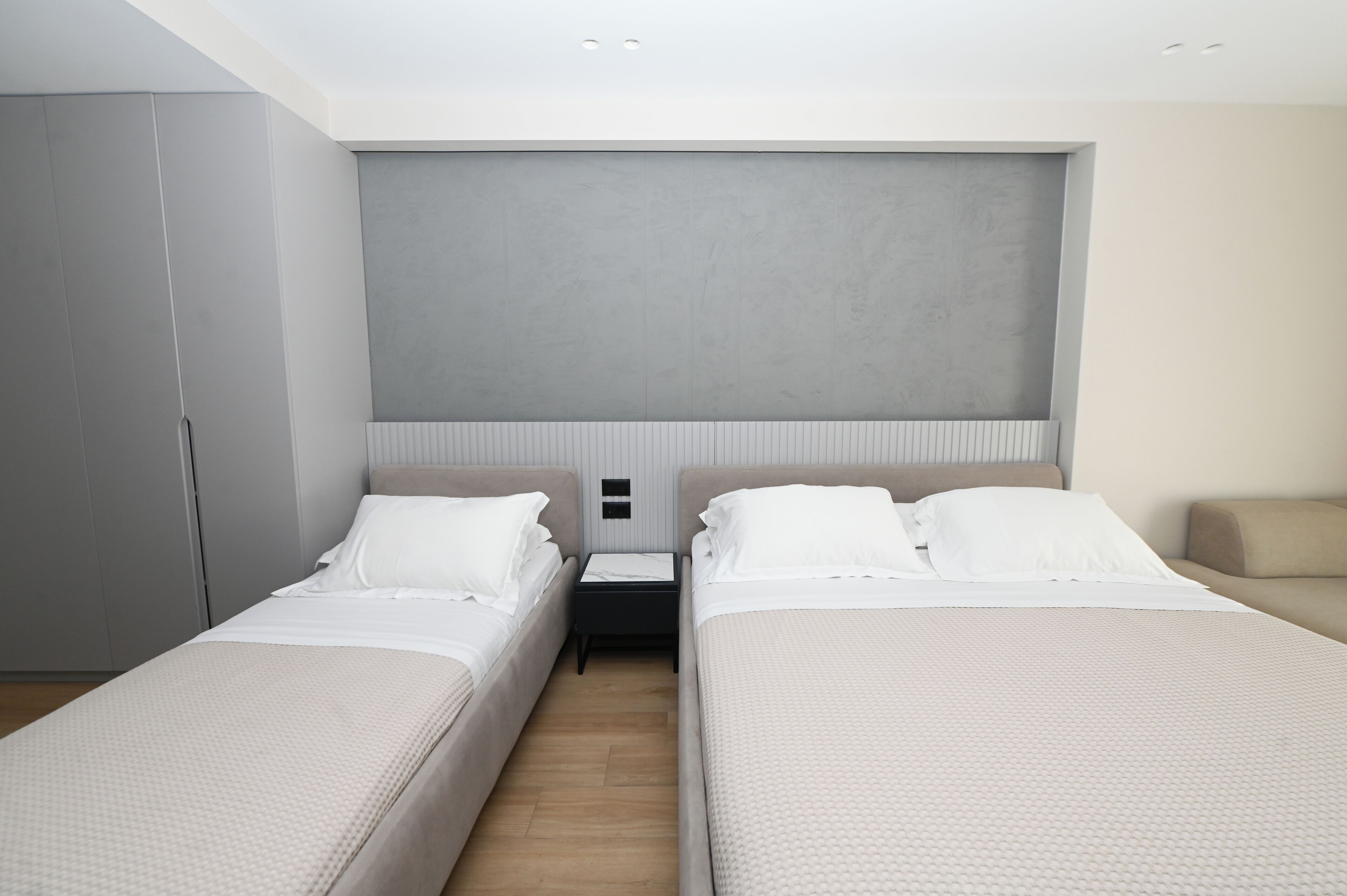 Habitación de hotel moderna con dos camas individuales, un cabecero grande de color gris, un armario y una mesita de noche.