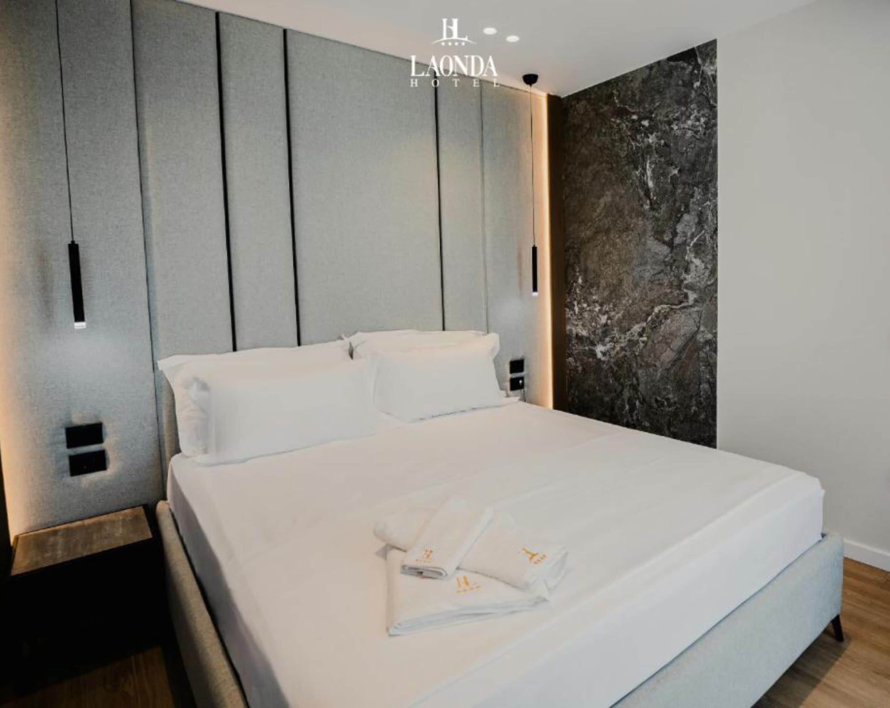 A modern hotel room with a large bed, grey panel headboard, and a marble accent wall.
