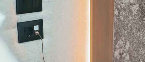 Una habitación de hotel con un teléfono sobre una mesita de noche, un enchufe y una pared con textura.