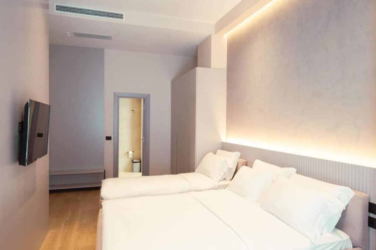 Habitación de hotel con dos camas, un televisor montado en la pared y un pasillo que conduce a un baño.