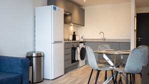A compact kitchen with a refrigerator, oven, and sink. There is a dining table with chairs and a trash bin. - GuestReady - Lux getaway in Liverpool (Liverpool)