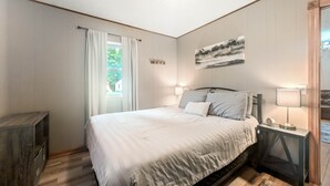 A bedroom with a bed, bedside lamps, a painting, and a window with curtains. - The Loon at Darling Shores (Alexandria)