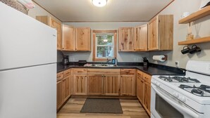 A compact kitchen with wooden cabinets, a white refrigerator, a stove, and a window with a view of greenery. - The Loon at Darling Shores (Alexandria)