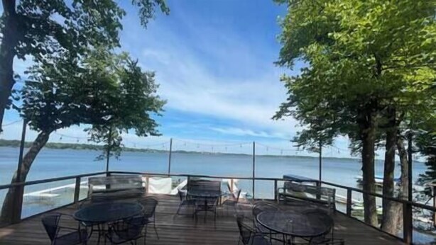 A deck with tables and chairs overlooking a lake with a bridge in the distance. - The Loon at Darling Shores (Alexandria)