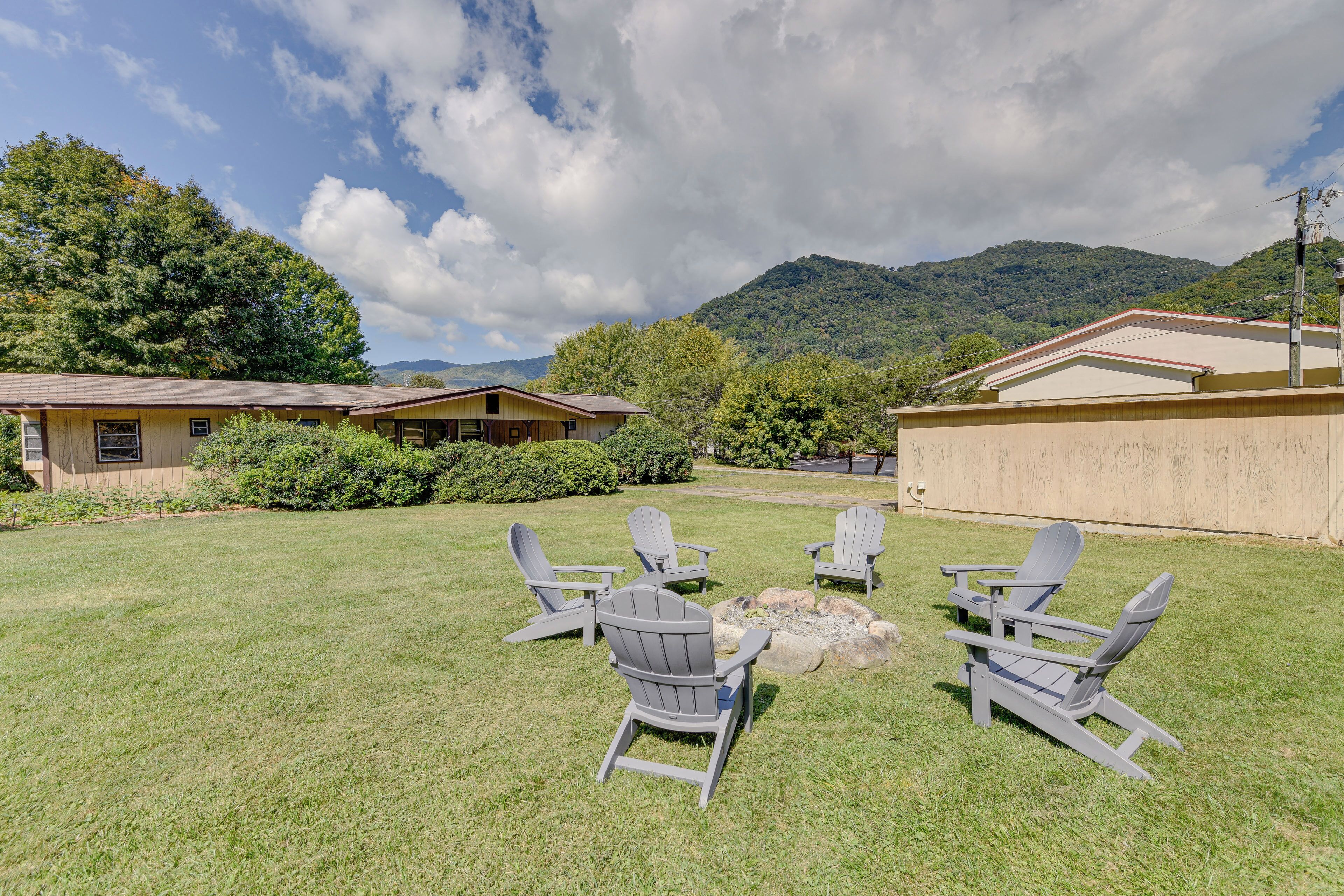 A backyard with Adirondack chairs arranged around a fire pit, a house with a red roof, and a mountainous backdrop.