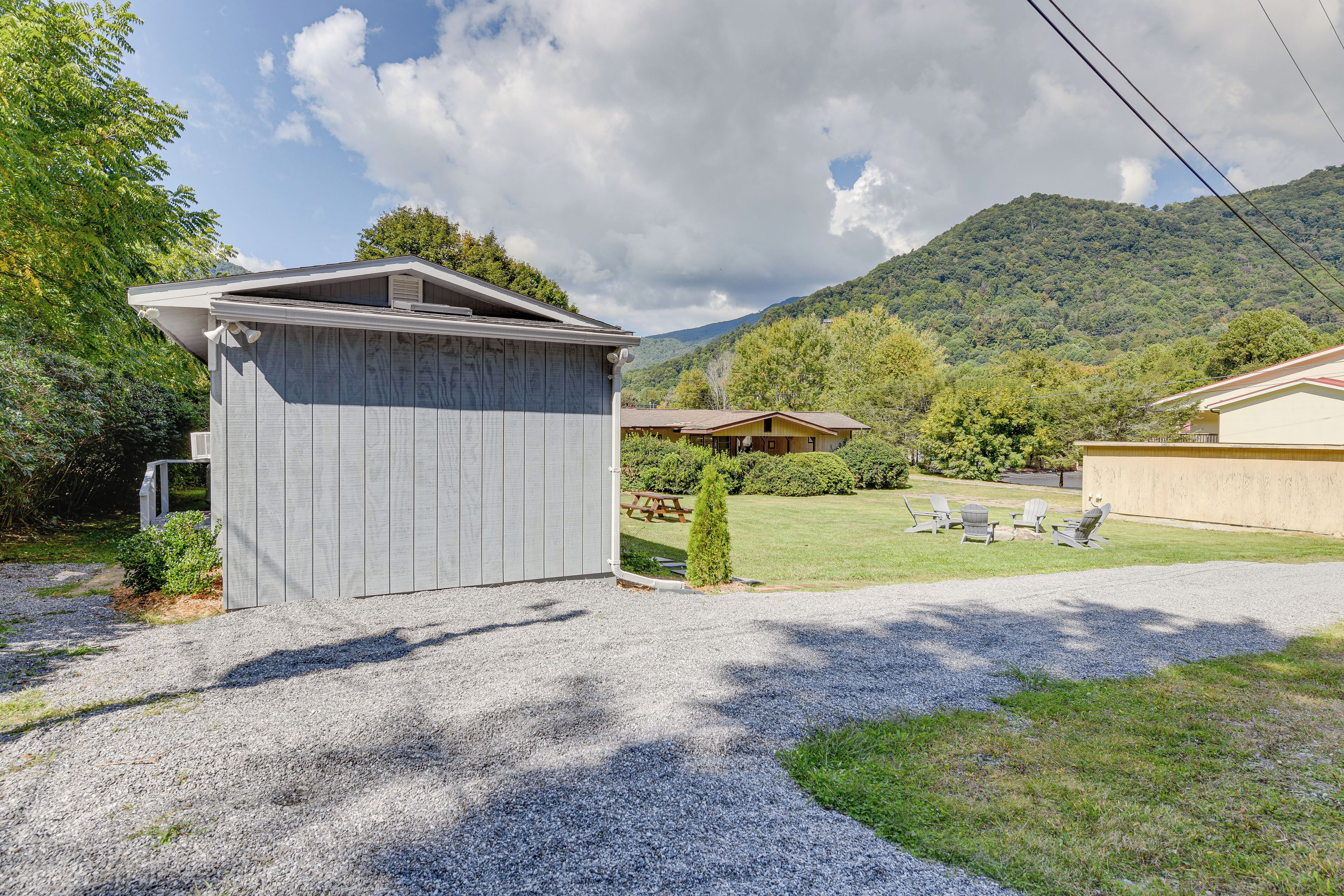 A wooden shed in a backyard with a gravel driveway, surrounded by greenery and a mountain in the background.