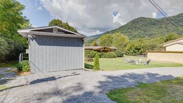 A wooden shed in a backyard with a gravel driveway, surrounded by greenery and a mountain in the background.