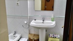 A bathroom with a white sink, a bidet, a small trash bin, a wicker basket, and a stool. - Loft Amplio con Garaje en Calle Rey Aurelio en Cangas de Onís (Cangas de Onís)