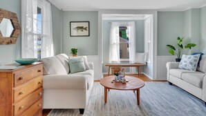 A living room with a beige sofa, a wooden coffee table, and a dining area with a small table and chairs. There is a large window with sheer curtains, a framed fish picture on the wall, and a potted plant. - Adorable Cottage with Cottage porch, walk to water & town (Westbrook)