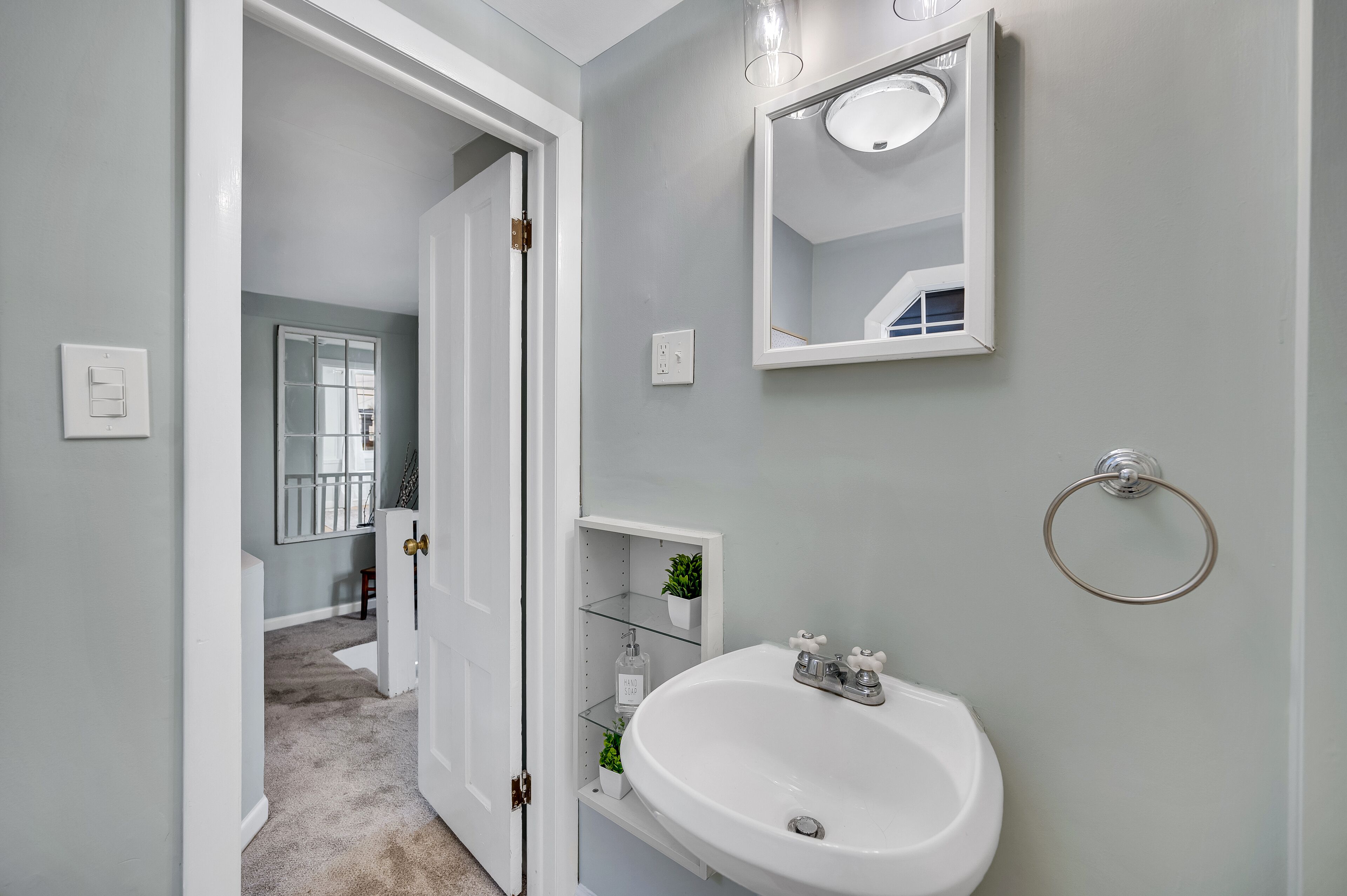 Un baño con un lavamanos blanco, un espejo y un aro para toallas. Hay una puerta que da a otra habitación.