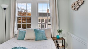 A bedroom with a bed, a window with curtains, a lamp, a small table with a plant, and a wall decoration. - Adorable Cottage with Cottage porch, walk to water & town (Westbrook)