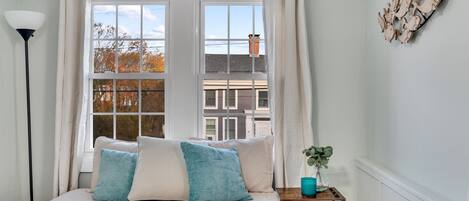 A bedroom with a bed, a window with curtains, a lamp, a small table with a plant, and a wall decoration.