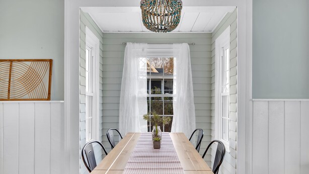 A dining area with a wooden table, white chairs, a checkered tablecloth, a vase with flowers, and a decorative ceiling light. - Adorable Cottage with Cottage porch, walk to water & town (Westbrook)