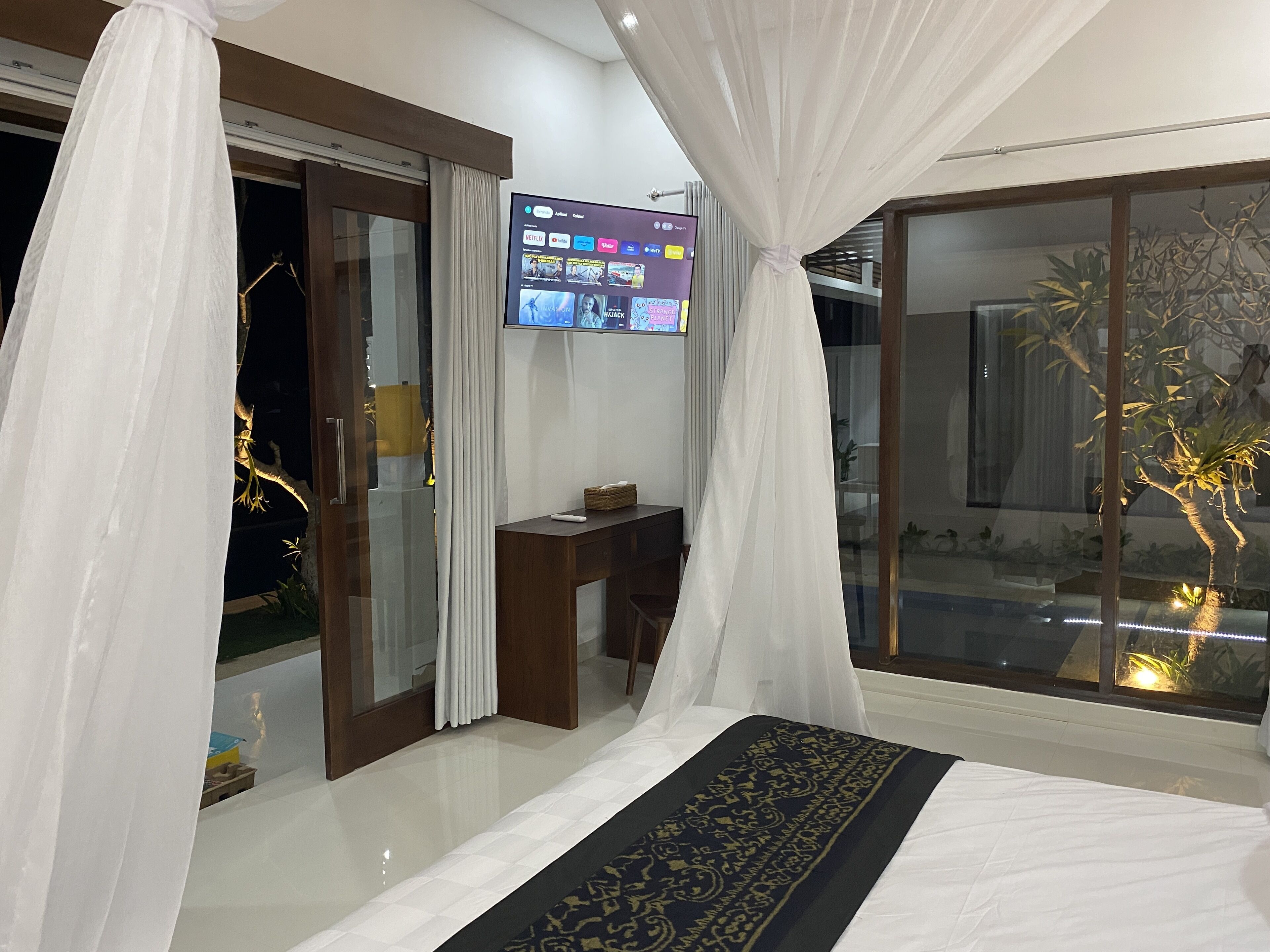 Un dormitorio con cama, televisor, escritorio y puerta corrediza de vidrio que da a un balcón con plantas.