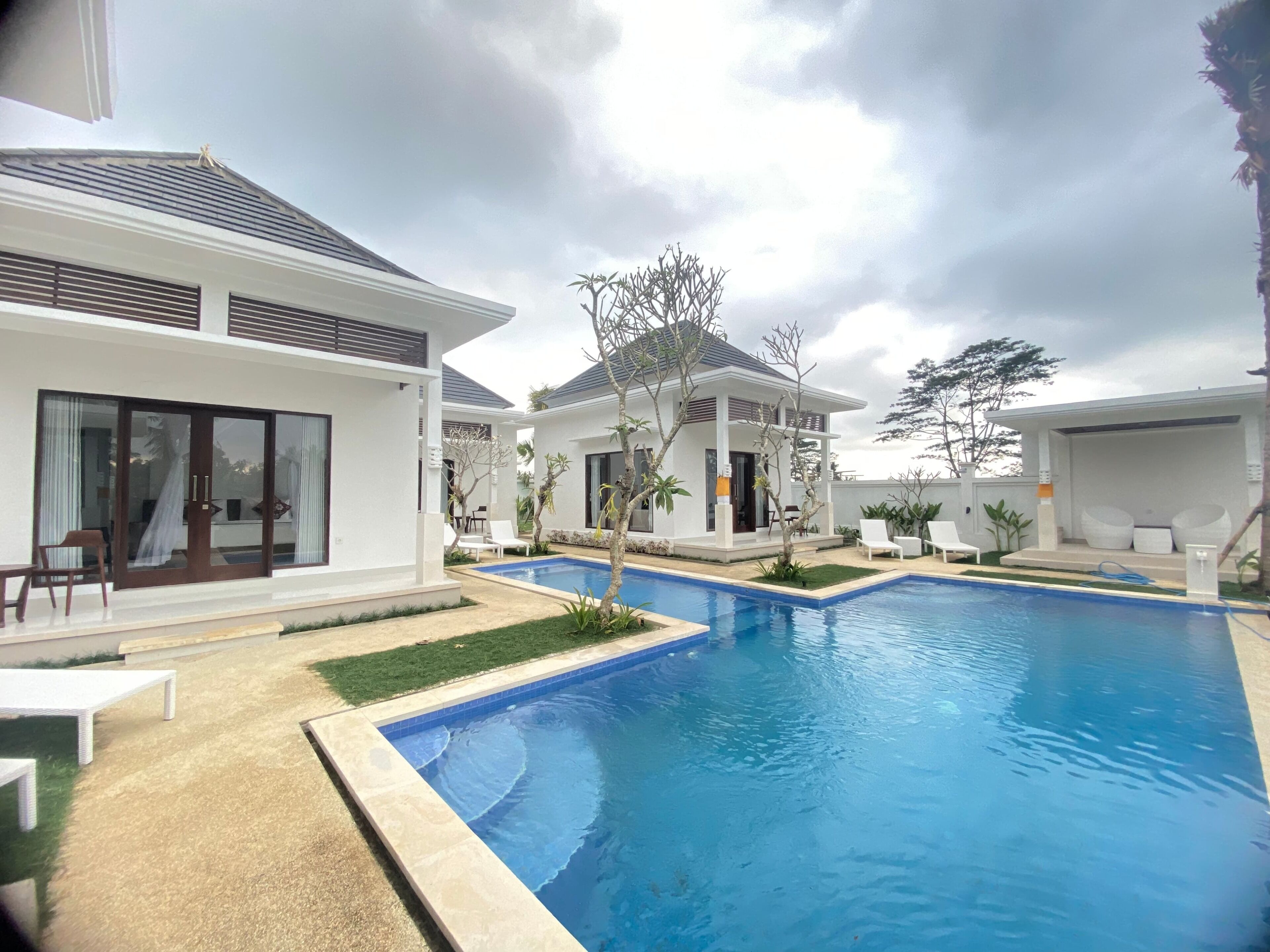 A modern pool area with a clear blue pool, surrounded by white buildings with large windows and doors, and a few trees.