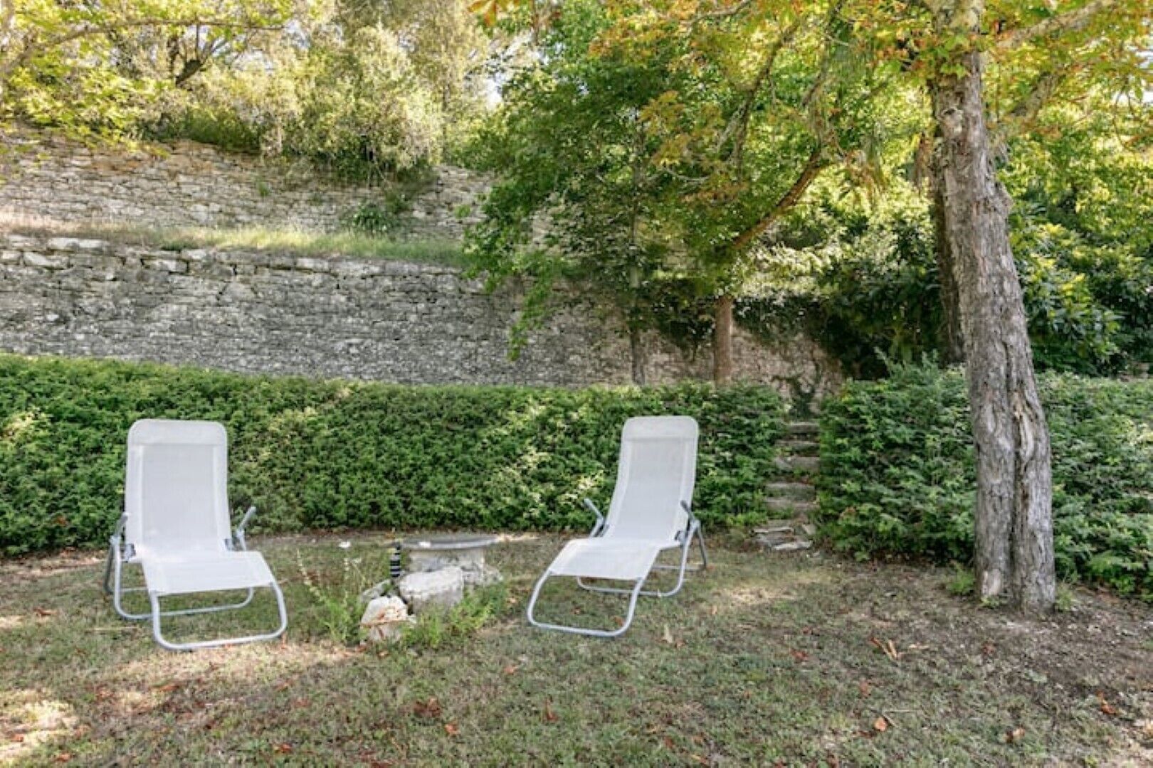 Deux fauteuils de jardin blancs, placés dans un jardin avec un mur de pierre et de la verdure en arrière-plan.