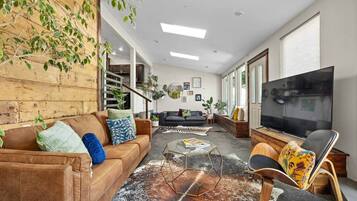 Una sala de estar con un sofá marrón, una mesa de centro de vidrio, un sofá negro, un televisor y una silla de madera.
